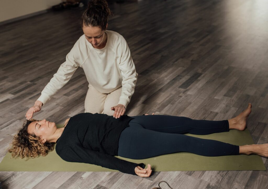 Eine Frau liegt auf einer Yogamatte, eine andere Frau unterstützt sie enegertisch beim Prozess