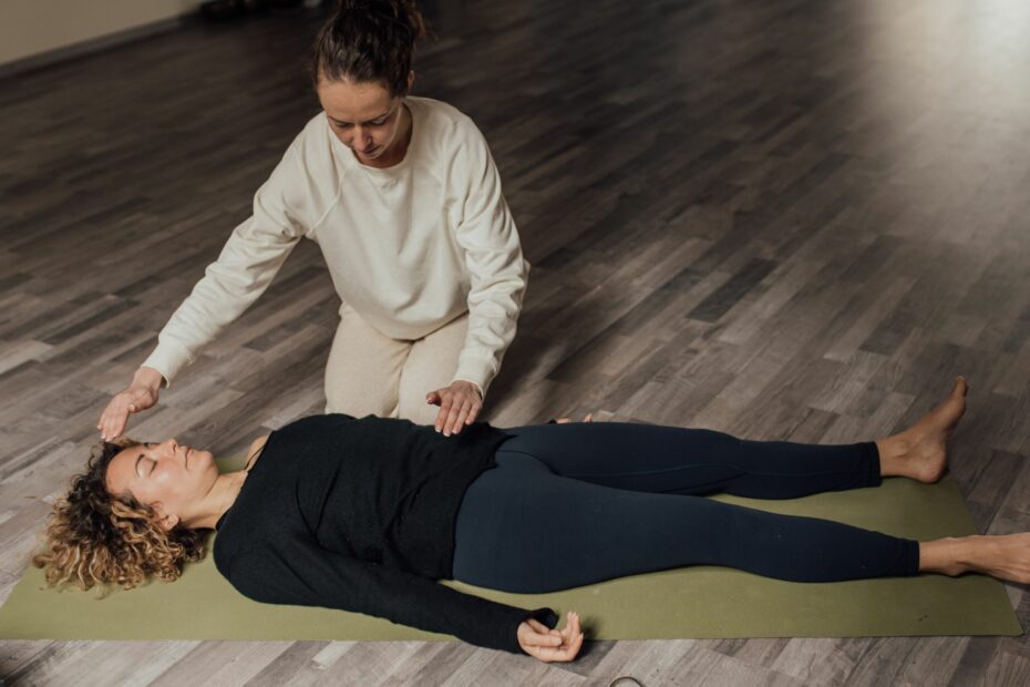 Eine Frau liegt auf einer Yogamatte, eine andere Frau unterstützt sie enegertisch beim Prozess