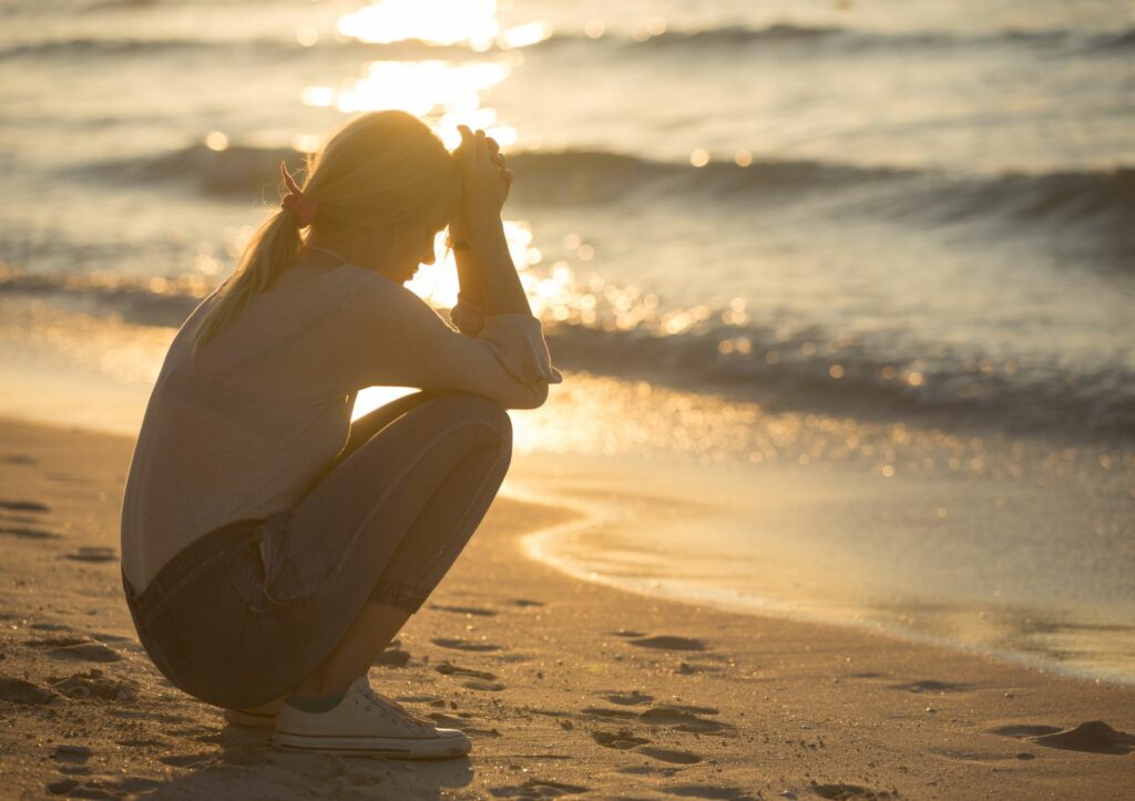 Eine Frau hockt am Strand und hält den Kopf in die Hände gestützt